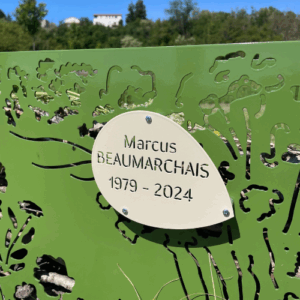 Photographie d'une plaque nominative de couleur ivoire sur une stèle cimélo de couleur verte dans un cimetière